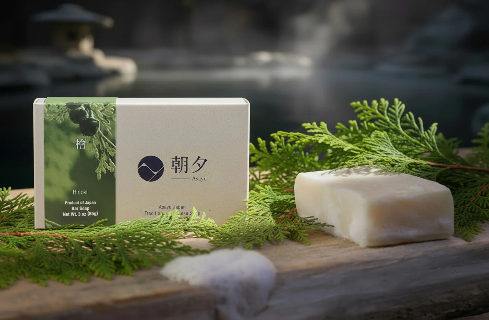 Two bars of soap on a wooden surface with greenery and a branded box in the background.