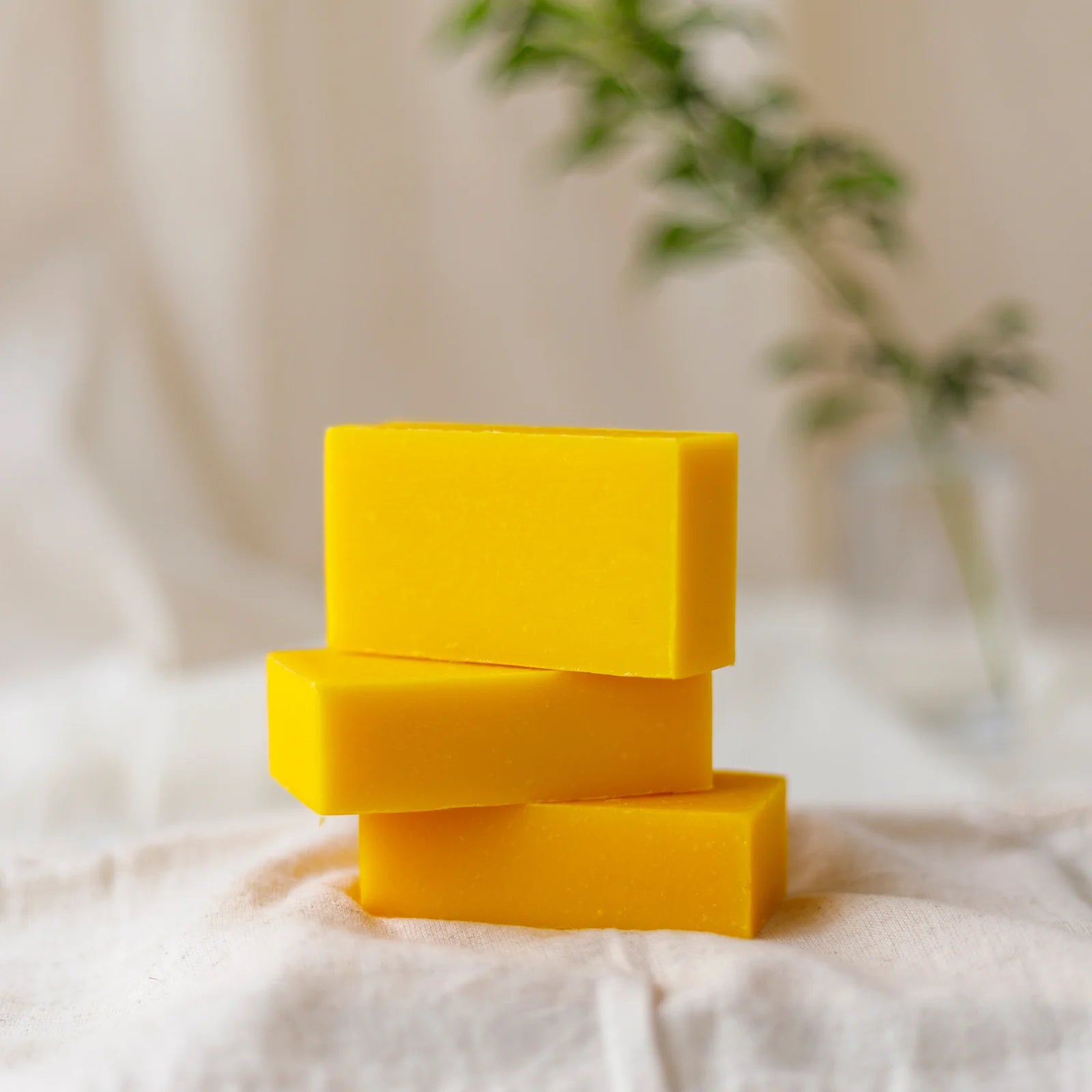 Three yellow soap bars stacked on a white fabric background with a blurred plant in the background.